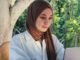 A woman in hijab using her laptop outside, surrounded by greenery and enjoying coffee.